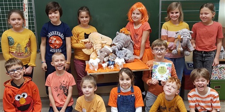 Powiększ grafikę: Dzieci z kl. IIa w pomarańczowych strojach z książką i pomarańczowymi słonikami.