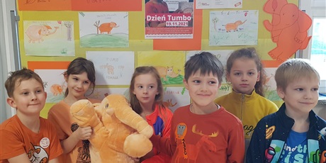 Powiększ grafikę: Dzieci z kl. Ib w pomarańczowych strojach z maskotką słonia na tle tablicy informującej o Dniu Tumbo.