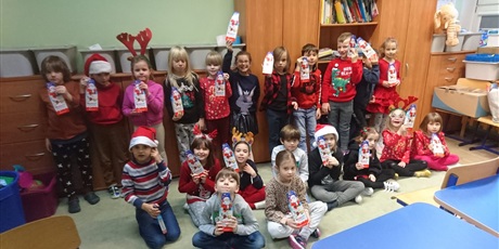 Powiększ grafikę: Uczniowie stoją w sali lekcyjnej ubrani w czerwone, mikołajkowe stroje i trzymają w rękach prezenty.