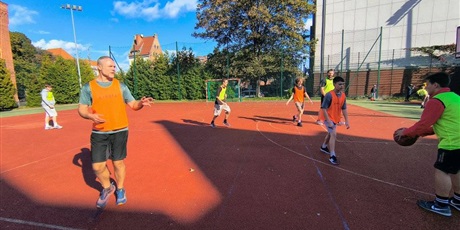 Powiększ grafikę: Uczniowie i rodzice rozgrywają mecz koszykówki na boisku szkolnym.