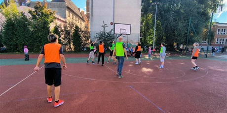 Powiększ grafikę: Uczniowie i rodzice rozgrywają mecz koszykówki na boisku szkolnym.
