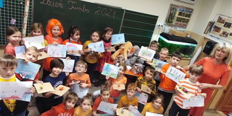 Powiększ grafikę: Dzieci z kl. IIa wraz z wychowawczynią w pomarańczowych strojach przedstawiają swoje prace plastyczne wykonane z okazji "Dnia Tumbo"
