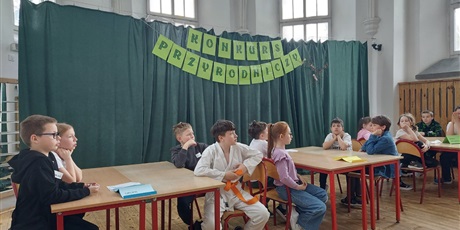 Powiększ grafikę: Przy trzech stolikach siedzą uczestnicy konkursu. W tle dekoracja z napisem.