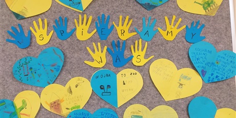 Powiększ grafikę: Dzieci wspierają Ukrainę. Na tablicy żółto-niebieskie serca z napisami przeciw wojnie. W środku rączki z napisem "Wspieramy Was".