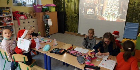 Powiększ grafikę: Dzieci w świetlicy szkolnej wykonują świąteczne ozdoby. Siedzą przy stolikach, na ścianie jest wyświetlone zdjęcie z przystrojoną choinką.