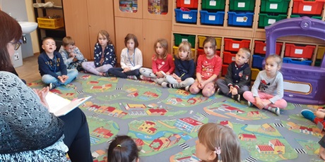 Powiększ grafikę: Dzieci z kl. 0a słuchają czytanej książki przez p. Mirkę z biblioteki. Dzieci siedzą na dywanie.
