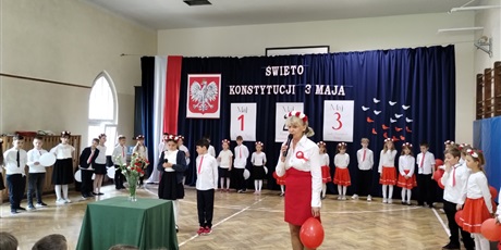 Powiększ grafikę: Uczniowie w odświętnych strojach stoją na tle dekoracji z napisem Święto Konstytucji 3 Maja., Nauczycielka zapowiada występ.