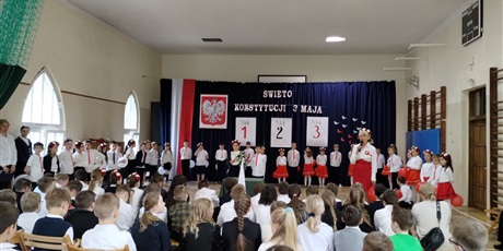 Powiększ grafikę: Uczniowie w odświętnych strojach stoją na tle dekoracji z napisem Święto Konstytucji 3 Maja.,