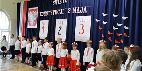 Powiększ grafikę: Uczniowie w odświętnych strojach stoją na tle dekoracji z napisem Święto Konstytucji 3 Maja.,