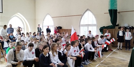 Powiększ grafikę: Sala gimnastyczna wypełniona uczniami w odświętnych strojach i biało-czerwonymi flagami.