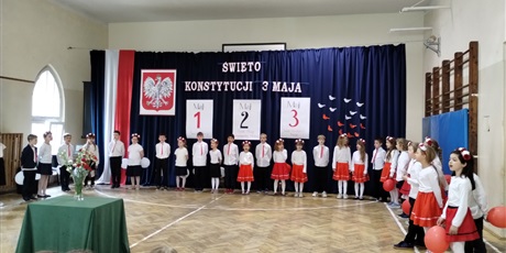 Powiększ grafikę: Uczniowie w odświętnych strojach stoją na tle dekoracji z napisem Święto Konstytucji 3 Maja.,