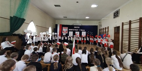 Powiększ grafikę: Uczniowie w odświętnych strojach stoją na tle dekoracji z napisem Święto Konstytucji 3 Maja., Dzieci trzymają w dłoniach balony w barwach narodowych.