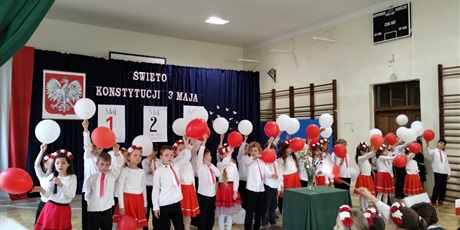 Powiększ grafikę: Uczniowie w odświętnych strojach stoją na tle dekoracji z napisem Święto Konstytucji 3 Maja., Dzieci trzymają w dłoniach balony w barwach narodowych.
