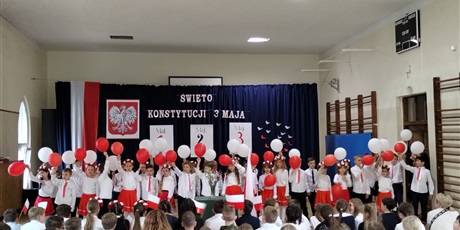 Powiększ grafikę: Uczniowie w odświętnych strojach stoją na tle dekoracji z napisem Święto Konstytucji 3 Maja., Dzieci trzymają w dłoniach balony w barwach narodowych.