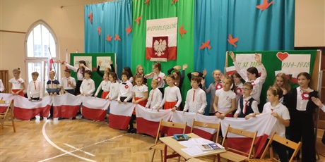 Powiększ grafikę: Na niebiesko-zielonym tle stoją uczniowie w strojach galowych z kotylionami. Uczniowie trzymają flagi Polski.