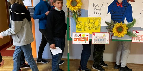 Powiększ grafikę: Uczniowie poszukują ukrytych w różnych zakamarkach szkoły kodów QR. Na korytarzu znajduje się tablica informująca o święcie szkoły i patronce.