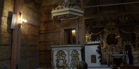 Powiększ grafikę: Zdjęcie ukazuje ambonę oraz kawałek drewnianej ściany. Ambona z wystroju jest podobna do ołtarza. Jest umieszczona nie wysoko. Obok znajduje się konfesjonał. 