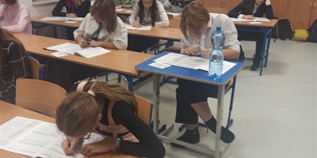 Powiększ grafikę: uczniowie ubrani w białe bluzki siedzą przy stolikach, piszą egzamin